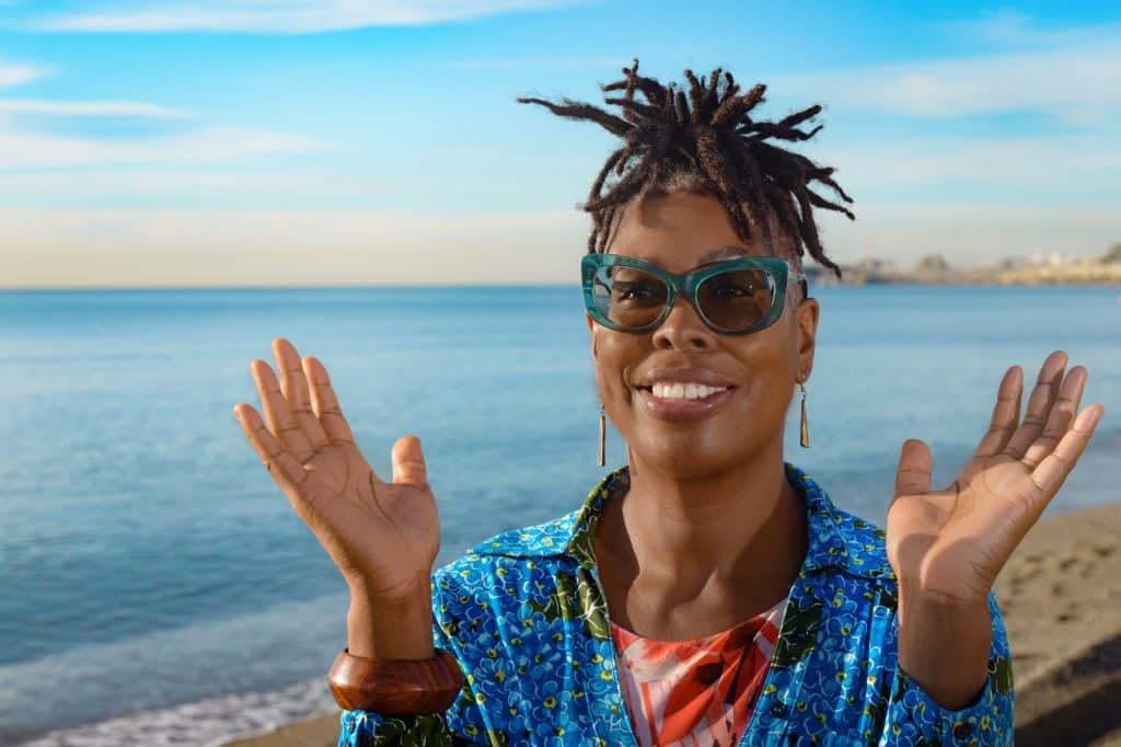 Image of Marcia Bravo Celebrant standing on the Malagueta beach posing for a photo smiling with hand wide open.  Sand and sea can be seen in the background of image.