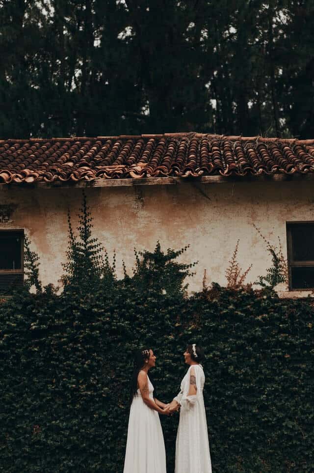 Image of two brides in their wedding dresses holding hands laughing together in front of a Mediterranean house with spiralling foliage.