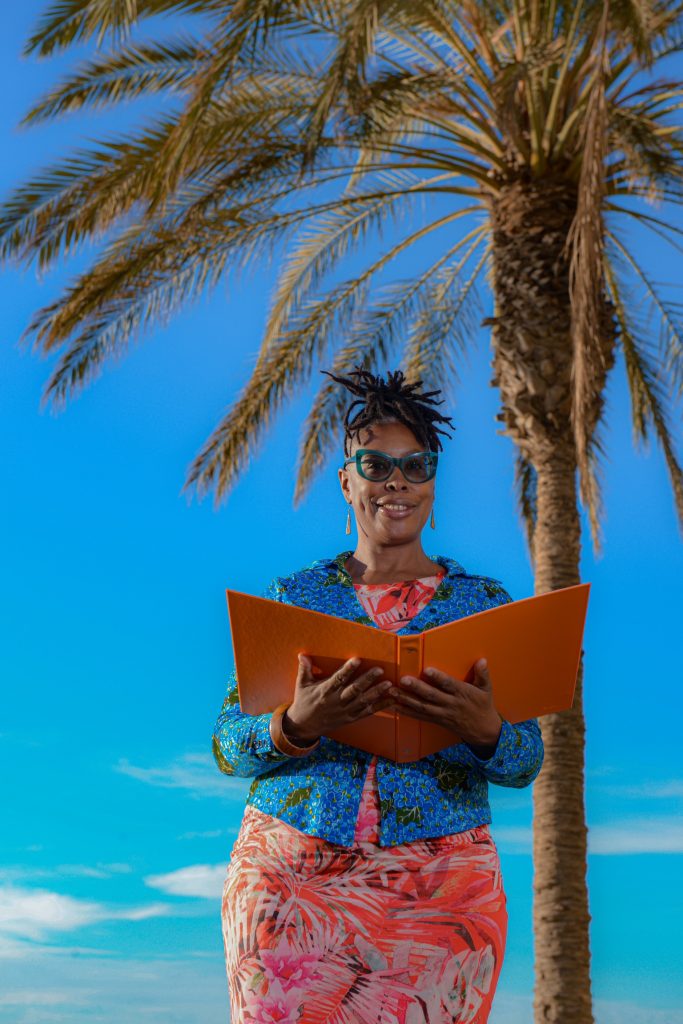 Image of Marcia Bravo Wedding Celebrant UK and Spain with palm tree in the background and blue sky.  Marcia is smiling whilst holding an orange folder in both hands.