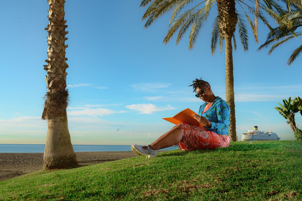 Image of wedding celebrant UK and Spain, Marcia Bravo. Marcia is seated on a grass verge next to the Malagueta beach writing.  In the distance is a large cruise ship.