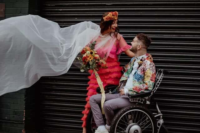 Why poems are excellent for a celebrant wedding. Image of wedding couple posing for photos in an urbanised location.  Both wear wedding attire with pinks, oranges and white. Bride stood at  husband´s side holding floral bouqet.  Husband is seated in a wheelchair.  Both look into each other eyes, smiling.