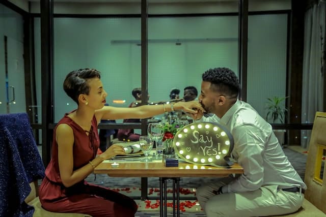 Image of a woman and man  sat at a table in a restaurant.  The woman is holding out her hand and the man appears to kiss her hand.  A sign with lights can be seen on the table with the words say yes and it appears it´s and engagement proposal.