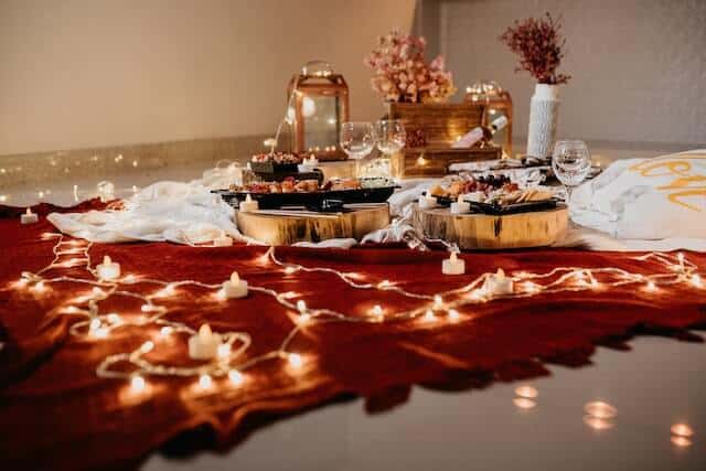 Image of red rug on floor adorned with electric tea/light candles, glass lantern, wine glasses and selection of snacks. Decorated with flowers possibly for a valenetine's proposal.