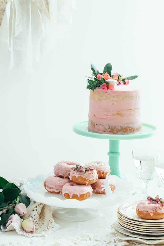 Image of wedding cake with pink icing and green foliage on a pale green cake tier with donuts with pink frosting below. Example of how wedding suppliers create magical moments.