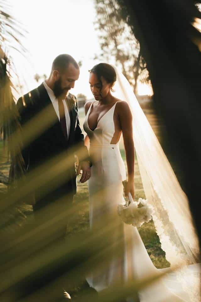 Image of a couple posing for a photo following their summer wedding in Spain. Bride and groom stand close together in outside space with green tropical foliage in background. Groom wears dark suit and bride long white dress with long trailing veil.