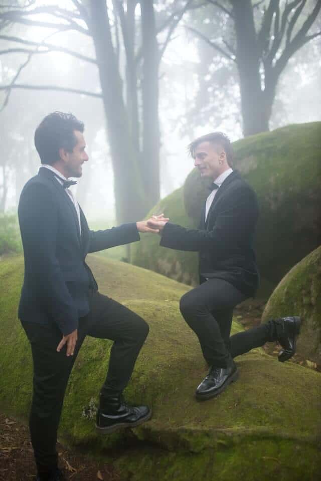 Going beyond the traditional in the way you share your vows gives your ceremony more meaning. Image of grooms on their wedding day outside on a raised lawn.  Both wear black suits and white shirts, one is on bent knees holding their partners ring finger.