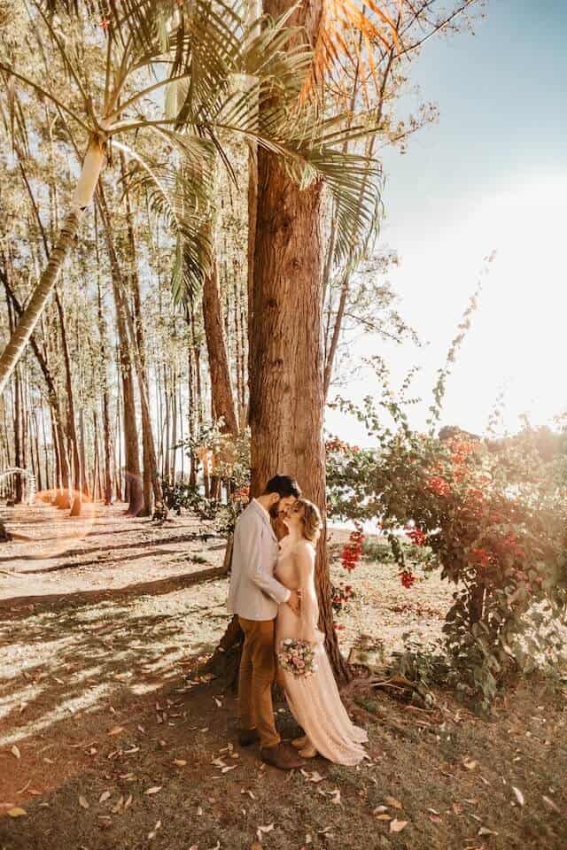 Image of man and woman in wedding attire in a forested zone in what could be an elopement planned on their own with a celebrant led wedding. man wears brown trousers and cream jacket.  Woman wears white lace dress and holds a bunch of flowersin one hand.