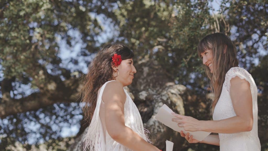 Image of couple smiling at their wedding for two. Both stand surrounded by nature below a grand oak tree. Both hold their vows.  Both wear white dresses with lace and fringes.