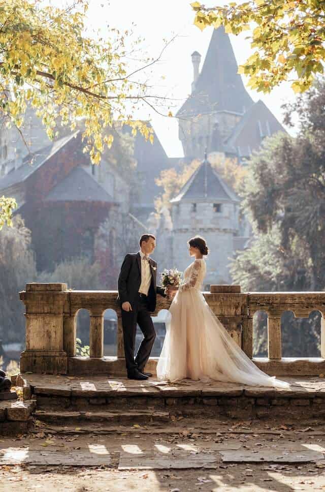 Image of a couple at their destination wedding . Groom wears a dark suit, white shirt and bow tie.  Bride full flowing white lace dress and holds mixed bouquet of flowers.  Couple appear to be standing near to an old stone bridge with a castle in the background.