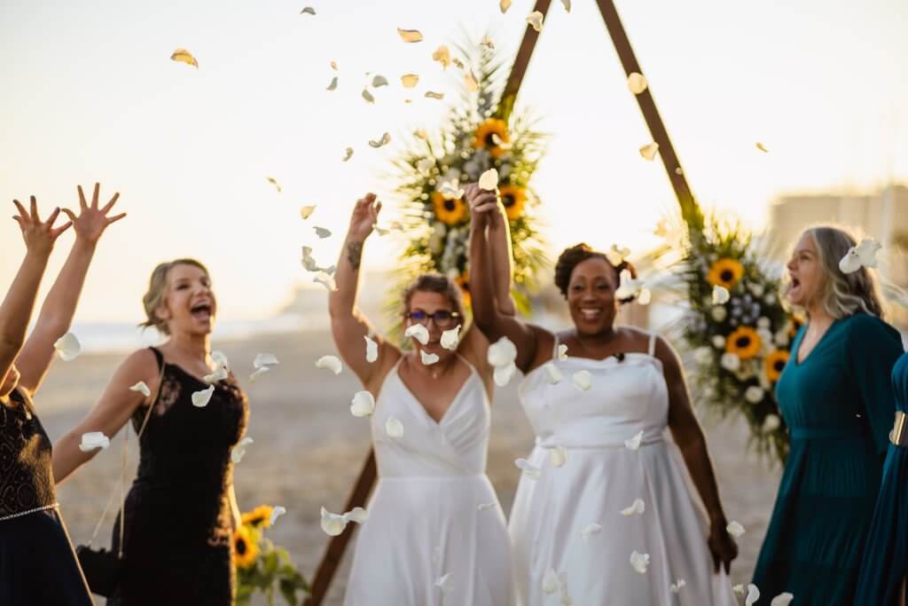 Image of a beach wedding ceremony, Couple exit their ceremony space smiling and holding one hand together raised high. Guests throw white confetti petals as they leave.  Both brides wear white flowing wedding gowns.