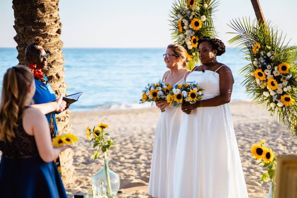 Image of same sex couple side by side on a Malaga beach, holding their sunflower wedding bouquet´s as they enjoy their blended family elopement ceremony officiated by Marcia Bravo Celebrant UK and Spain.  Both brides wear white flowing dresses with sunflower printed baseball boots.  Younger member of  their blended family looks on close by.