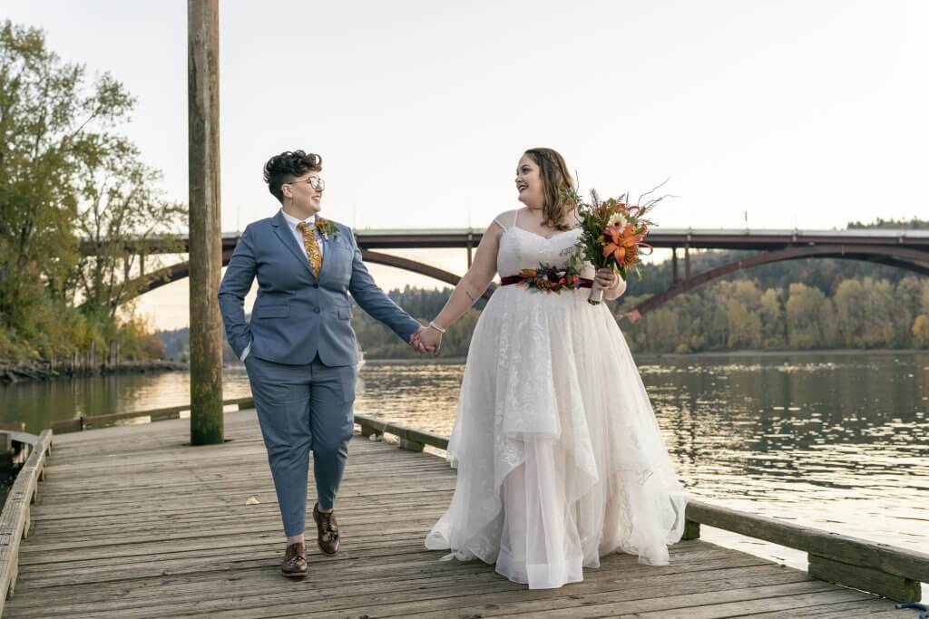 A joyful same sex couple hold hands and take a romantic sunset walk along a river after their off cruise intimate wedding ceremony.