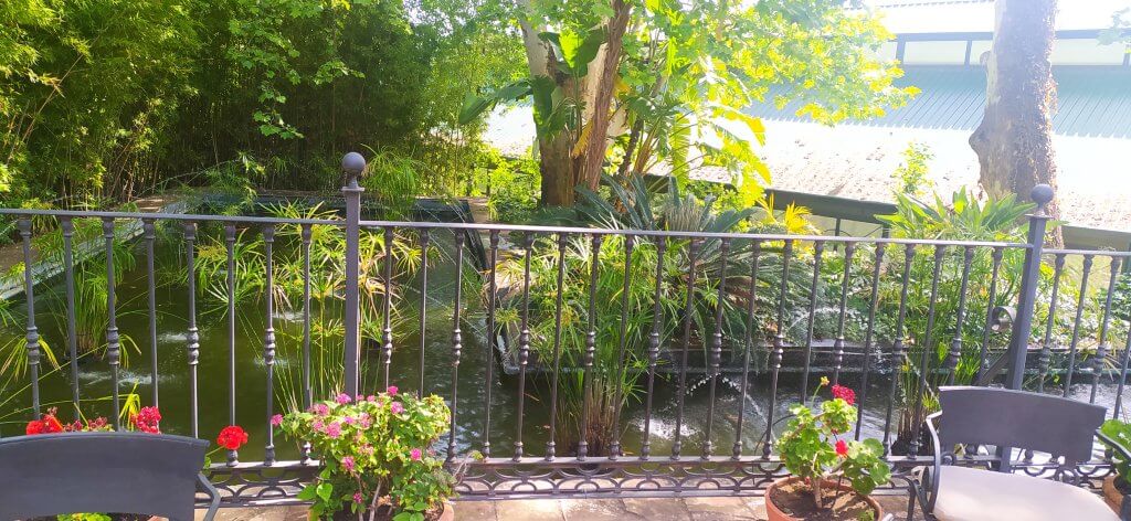 A peaceful garden scene with a pond, surrounded by lush greenery and plants, viewed from behind a decorative metal fence with flowers and patio chairs in the foreground.