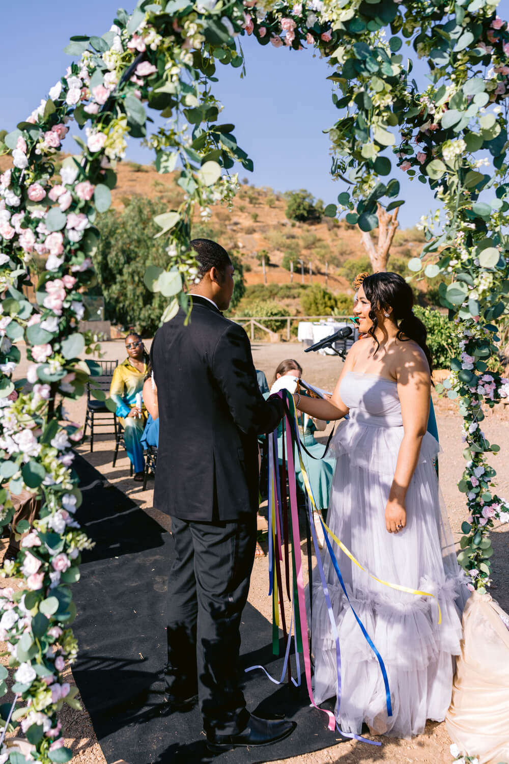 A bride and groom holding hands under a floral archway outdoors with guests in the background.