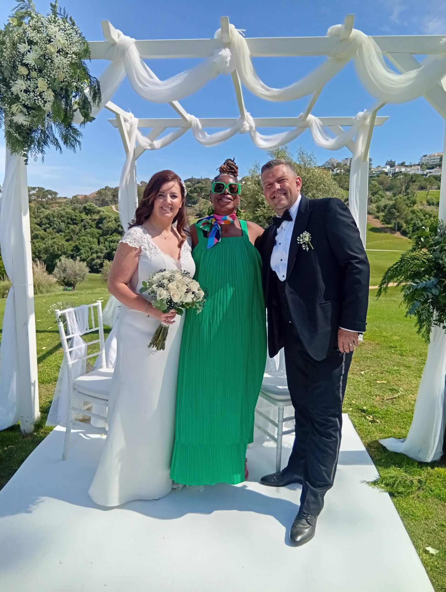 A joyful moment capturing a bride, a guest in a vibrant green dress, and a groom smiling under a decorated arch at an outdoor wedding ceremony.