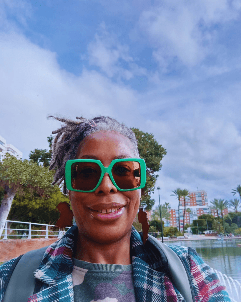A smiling individual with gray hair, wearing large green sunglasses and Africa-shaped earrings, poses outdoors near a scenic water feature and palm trees.