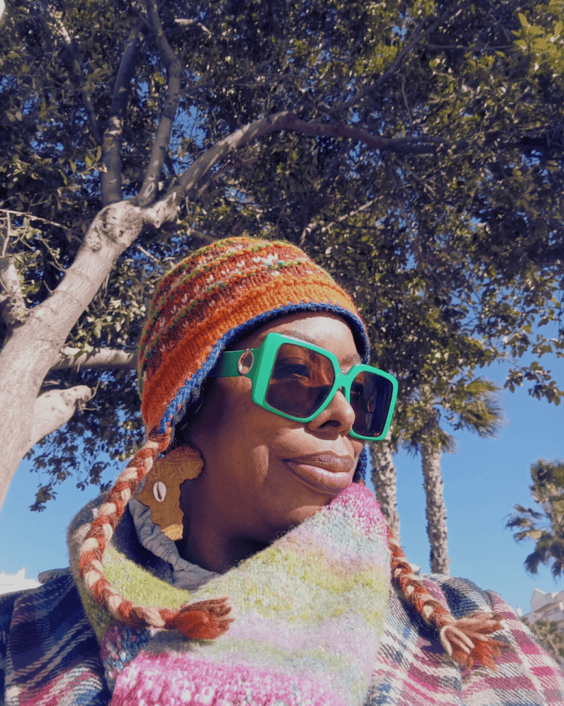 A woman wearing colorful attire, oversized green sunglasses, and a multi-colored scarf smiles against a backdrop of trees and palms.