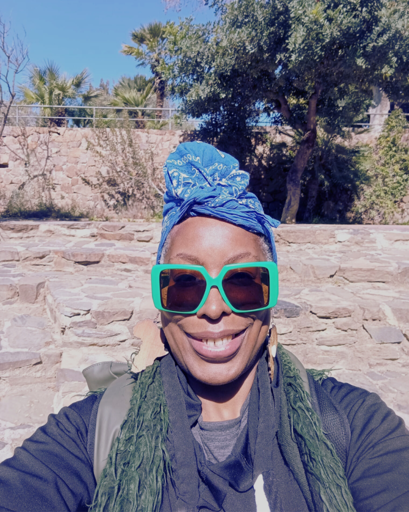 A smiling individual wearing oversized green sunglasses and a blue bandana, set against a backdrop of stone walls and greenery.