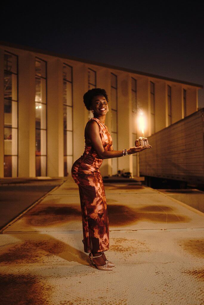 A woman in a shimmering brown dress holds a birthday cake with a sparkler, smiling in an outdoor setting at twilight.