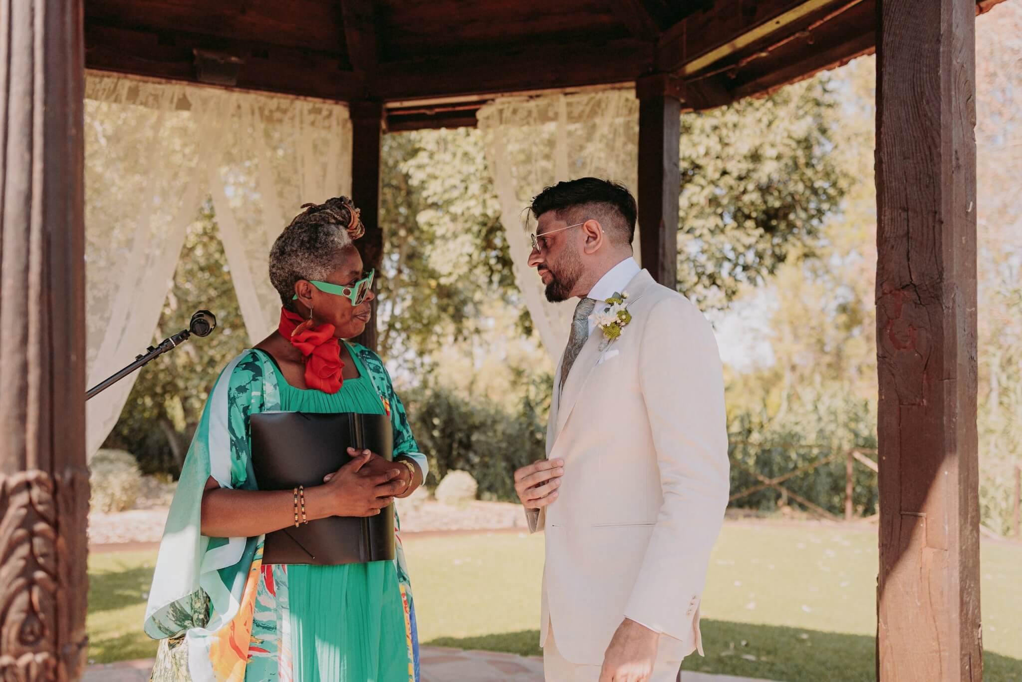 A couple engages in a heartfelt conversation under a gazebo, surrounded by natural greenery, with the officiant wearing vibrant colors and sunglasses.