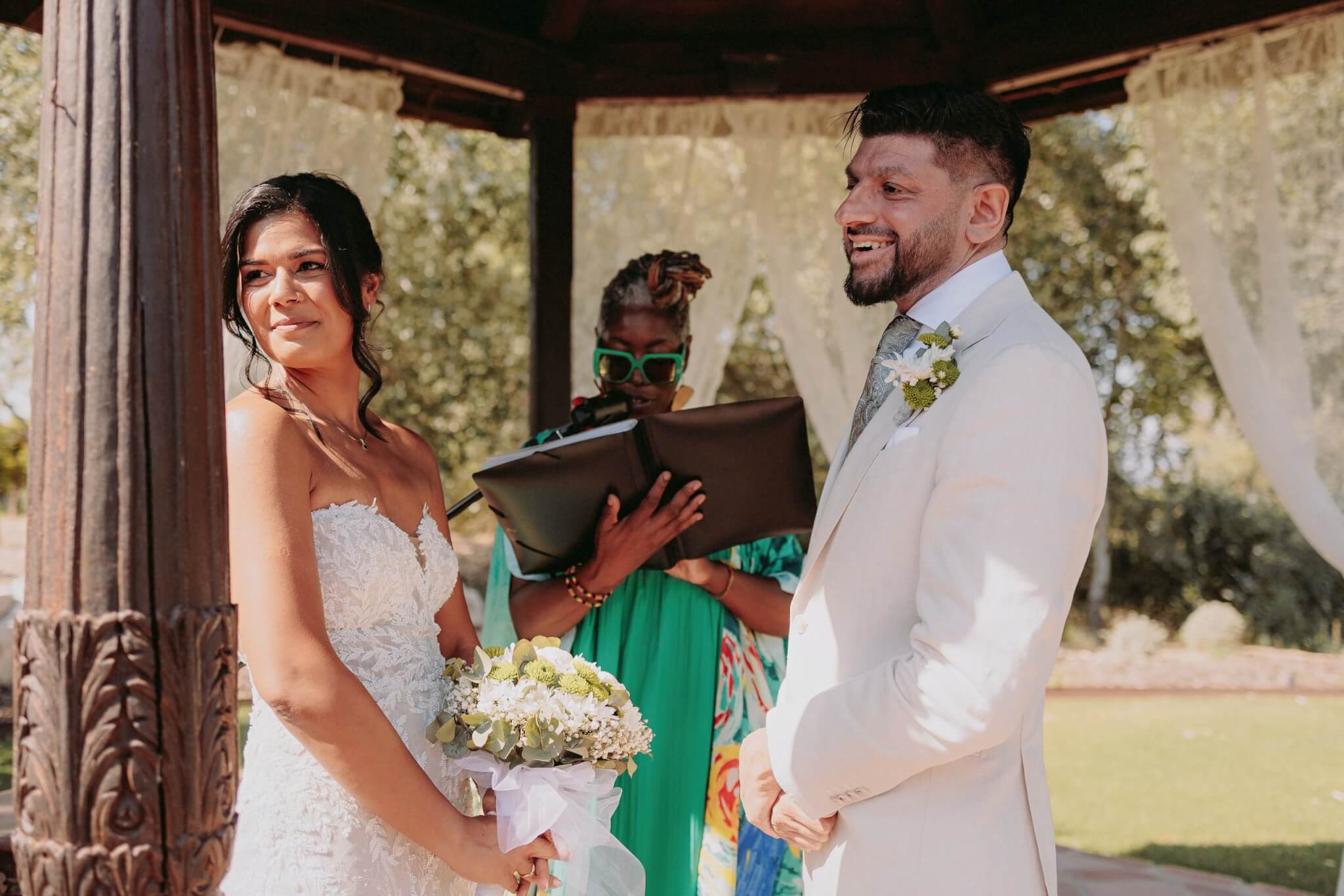 A bride and groom share a joyful moment during their wedding ceremony, standing under a gazebo with floral decorations, while a celebrant officiates the event.