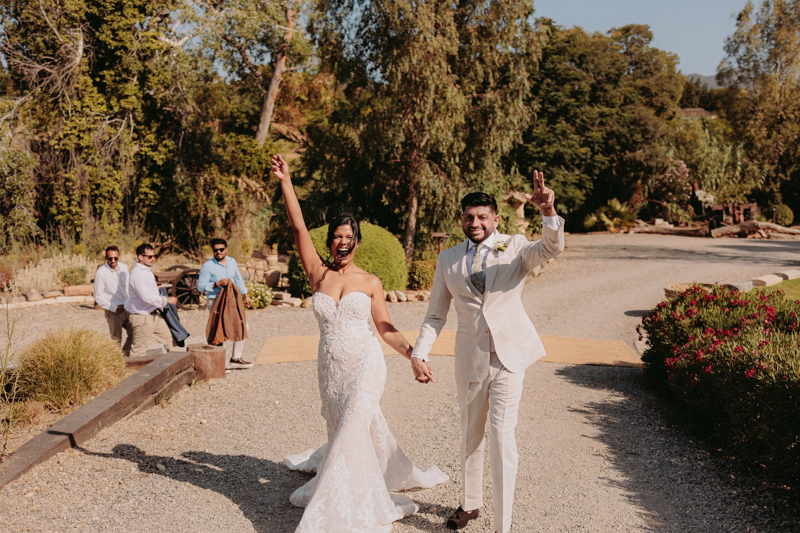 A bride and groom celebrate their wedding, walking hand in hand with joyful expressions, surrounded by family and a scenic outdoor setting.