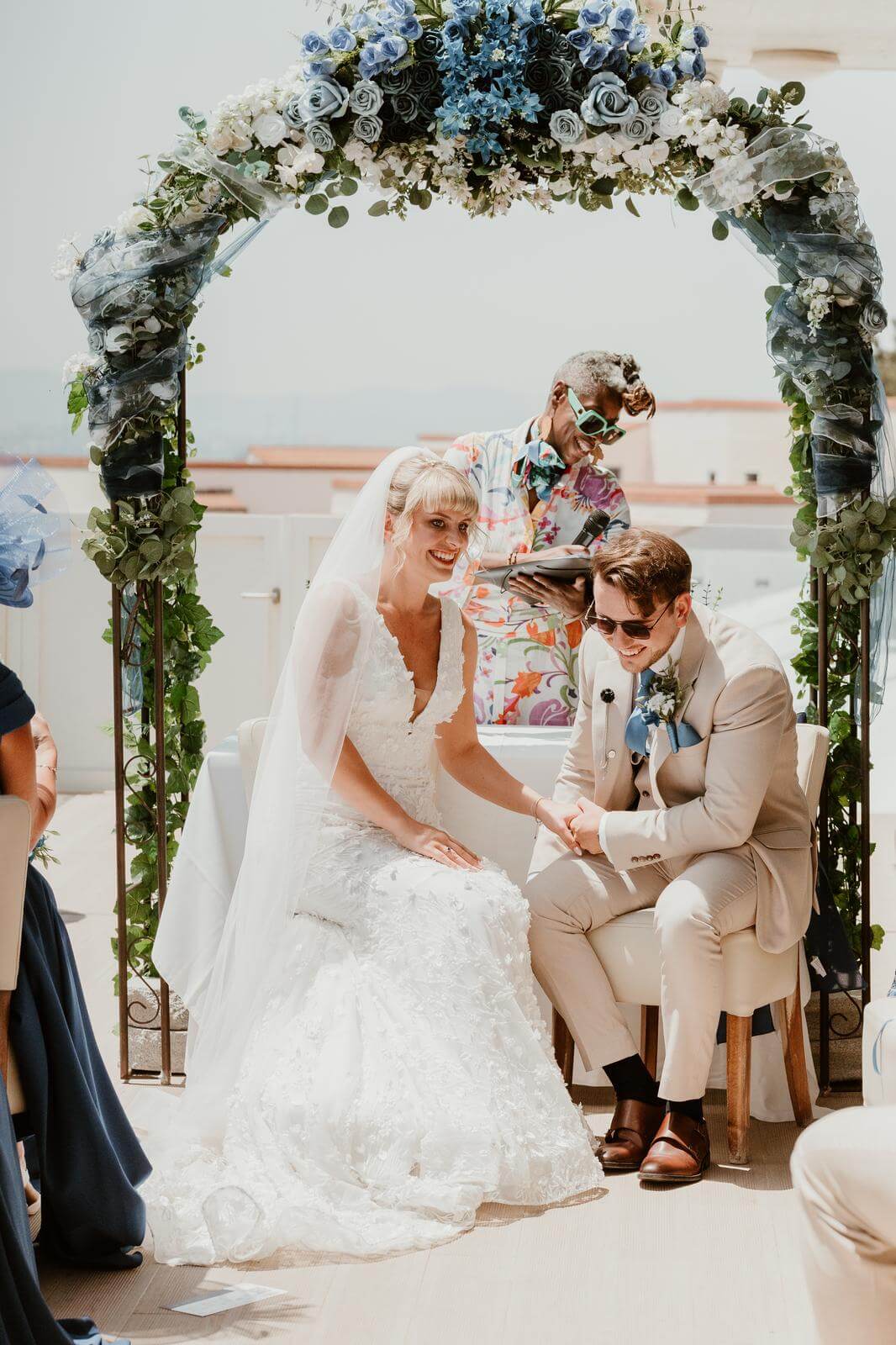 A joyful couple exchanging vows under a floral arch, with a lively officiant and guests in elegant attire.
