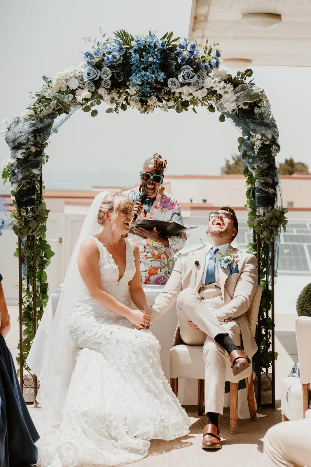 A bride and groom smiling happily during their wedding ceremony under a floral arch, with a celebrant speaking joyfully in the background.
