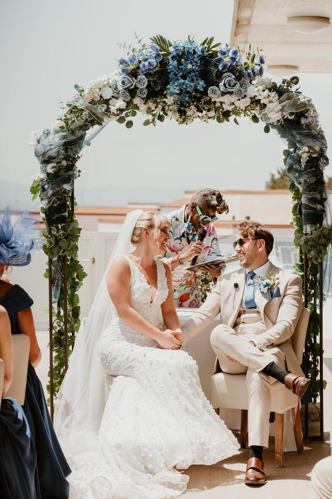 A couple smiles at each other during a wedding ceremony, seated beneath an ornate floral arch while the officiant prepares to conduct the ceremony.