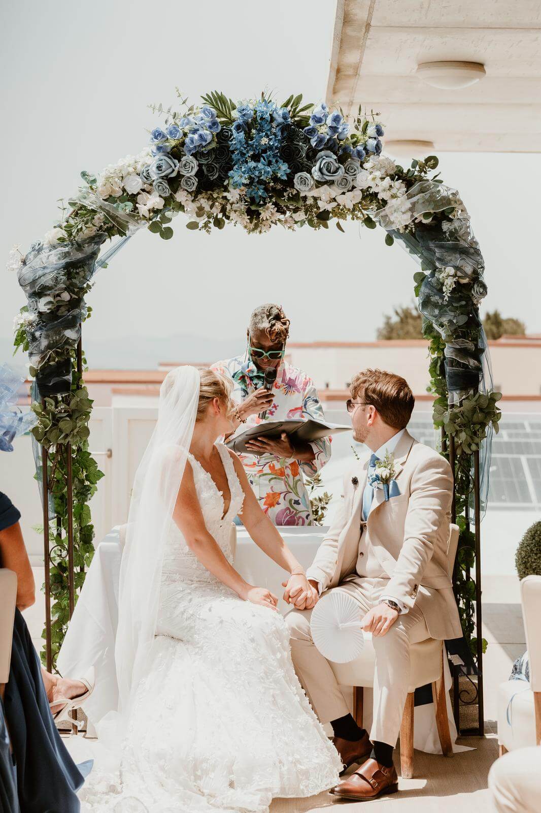 Bride and groom holding hands during their wedding ceremony beneath a decorated floral arch, with an officiant in a colorful outfit.