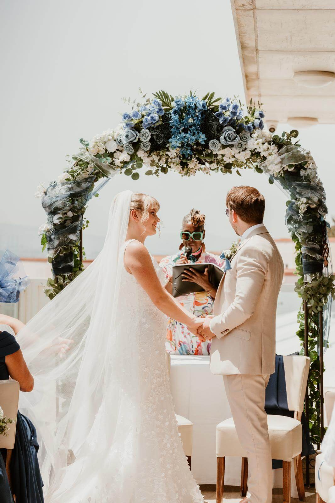 A bride and groom hold hands during their wedding ceremony, with a celebrant officiating under a beautifully decorated floral arch.
