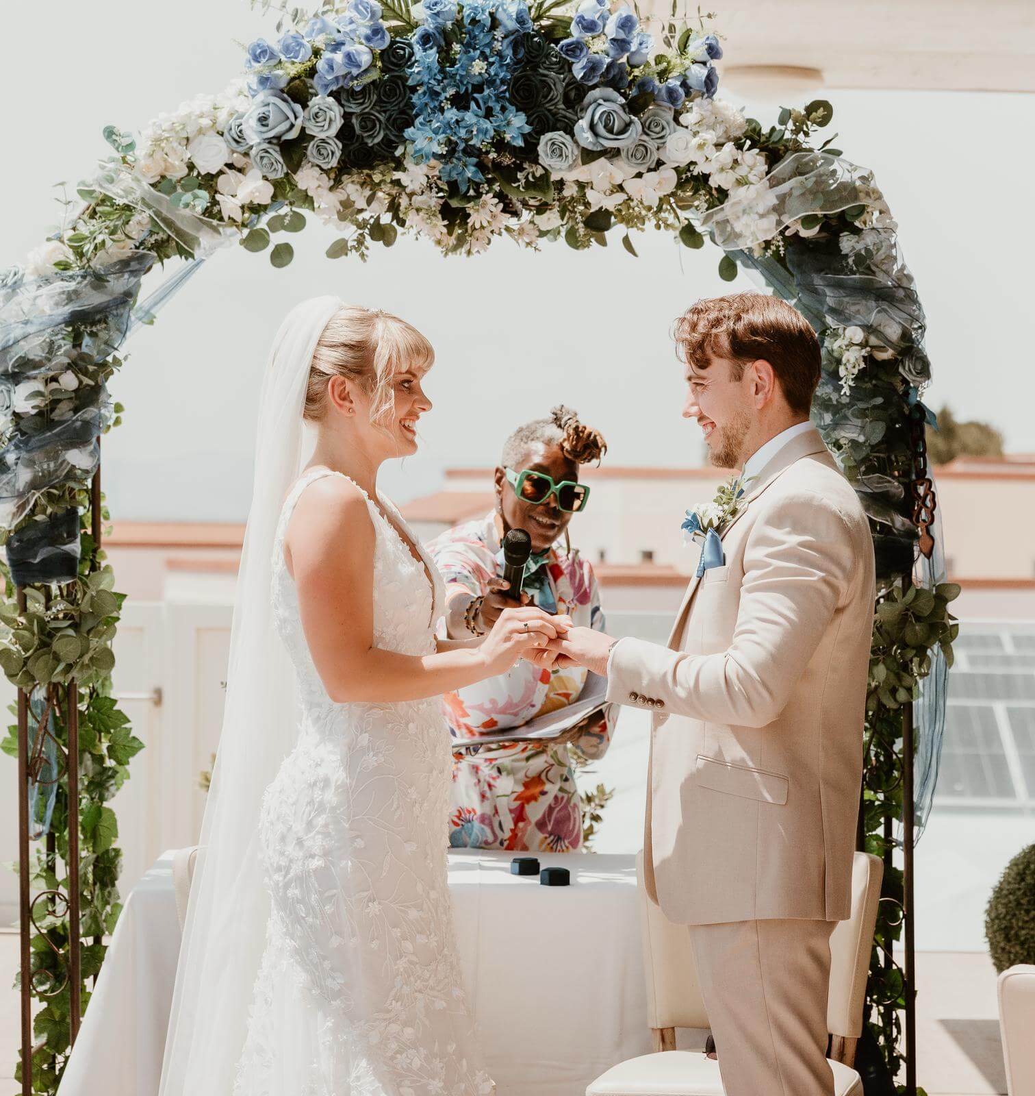 A bride and groom exchange rings during their wedding ceremony, with a celebrant officiating under a beautifully decorated floral arch.