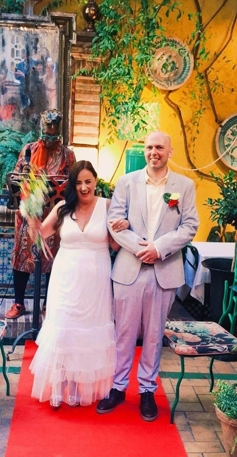 A joyful couple stands together on a red carpet during their wedding ceremony, with lush greenery and colorful decor in the background. The bride holds a bouquet and smiles brightly beside her groom.