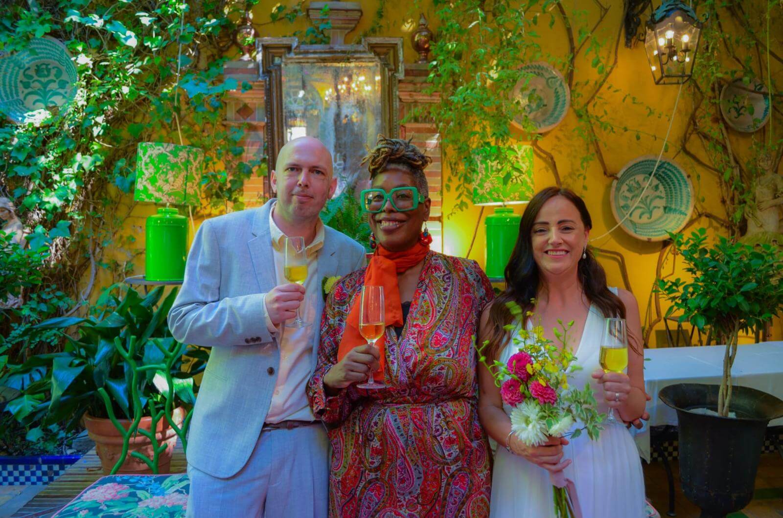 Three friends raise their glasses in a lively, colorful setting filled with plants and decorative elements.