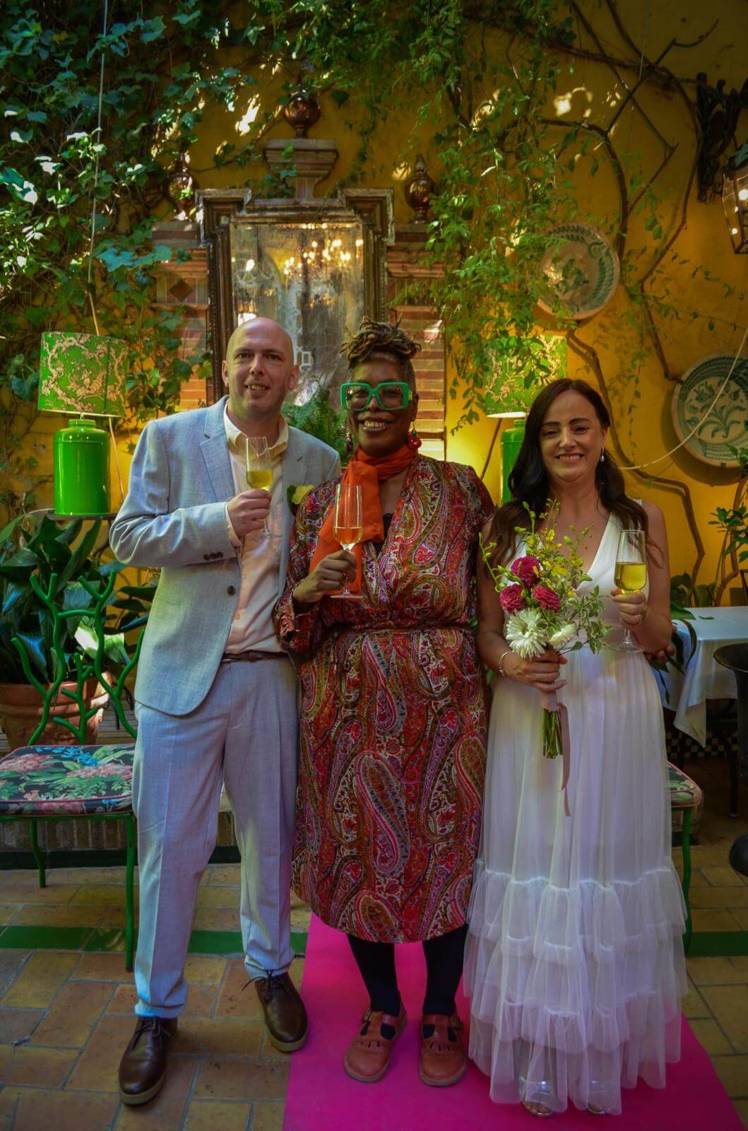 Three individuals in formal attire holding drinks, standing in a vibrant garden-like space with greenery and decorative elements in the background.