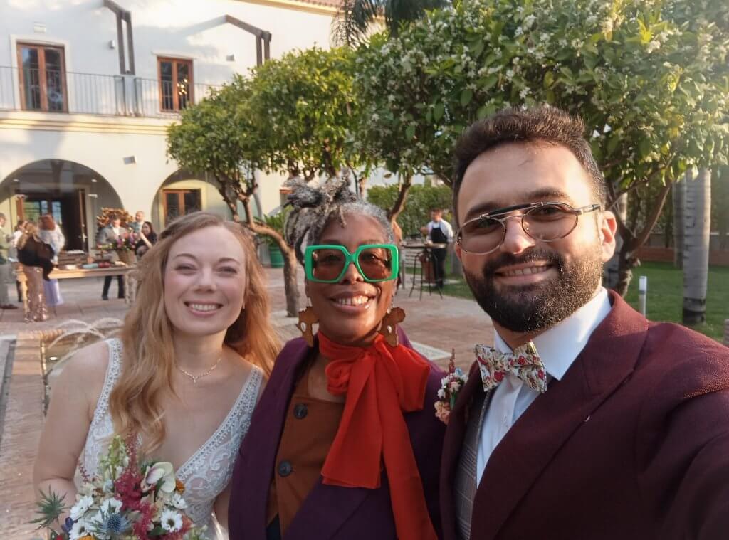 A smiling trio poses for a selfie at a wedding, with a lush garden and festive setting in the background.